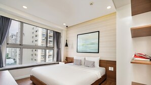 A modern hotel room with a large bed, a view of city buildings, a framed artwork on the wall, and a small shelf with books. - Scenic Valley Serviced Apartment (Ho Chi Minh City)