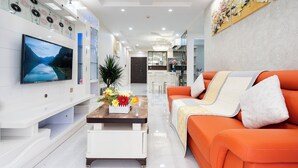 A modern living room with a flat-screen TV, a sofa, a coffee table with flowers, and a kitchen in the background. - Scenic Valley Serviced Apartment (Ho Chi Minh City)