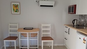Une cuisine compacte dotée de meubles blancs, d’un four à micro-ondes, d’une bouilloire et d’un petit réfrigérateur. On y trouve une table à manger avec deux chaises, ainsi qu’une chaise individuelle avec un plateau.