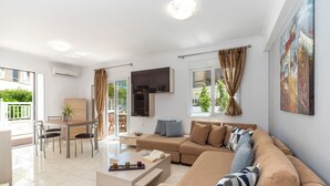 A modern living room with a sofa, coffee table, dining table, and chairs. There is a TV mounted on the wall and a painting above the sofa. - Haven Home: Central & Quiet- Elli Beach & Old Town (Rodos)