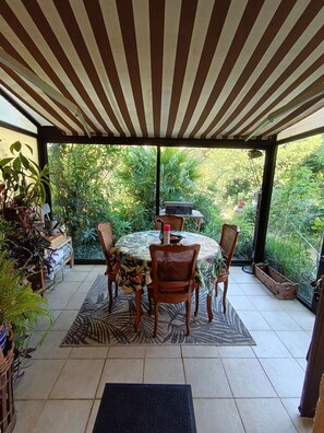 A covered patio with a round table and chairs, surrounded by potted plants and a view of greenery.
