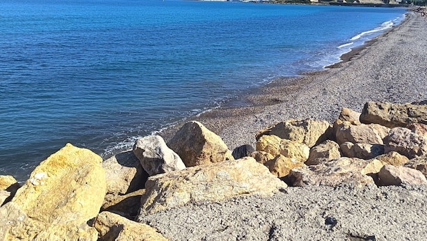 Une plage côtière avec des galets et des rochers, un ciel bleu dégagé et des bâtiments qui se dessinent à l’horizon.