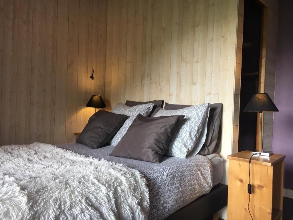 A bedroom with a wooden wall, a bed with grey bedding, two bedside lamps, and a wooden nightstand.