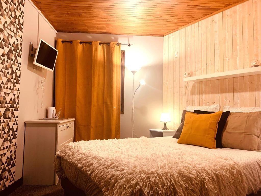 A bedroom with a wooden ceiling, a bed with a fluffy blanket, a bedside table with a lamp, and a TV on the wall.