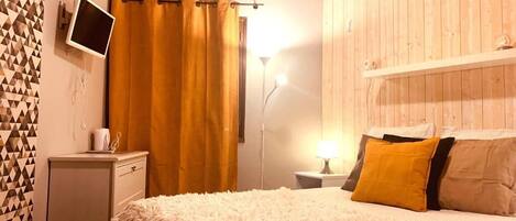 A bedroom with a wooden ceiling, a bed with a fluffy blanket, a bedside table with a lamp, and a TV on the wall.