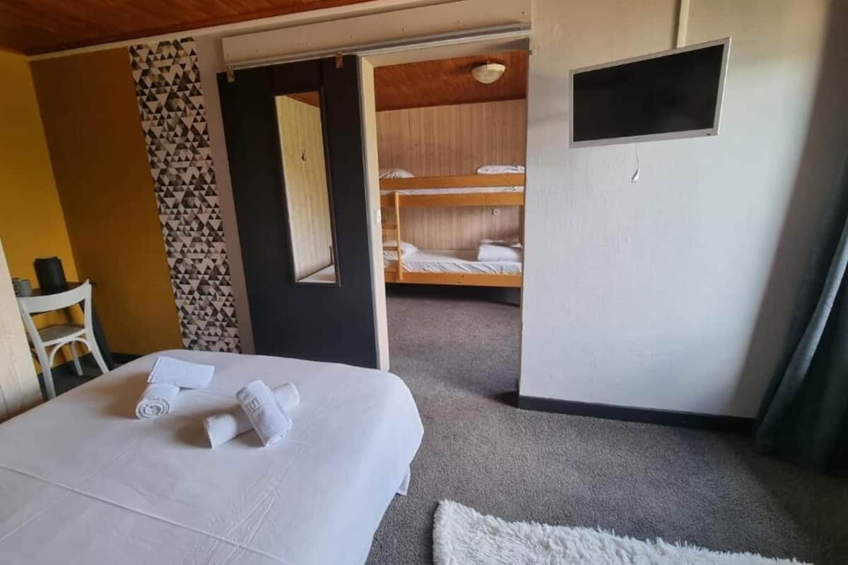 Habitación de hotel con cama, escritorio, silla, puerta que da a una zona con literas y un televisor pequeño.