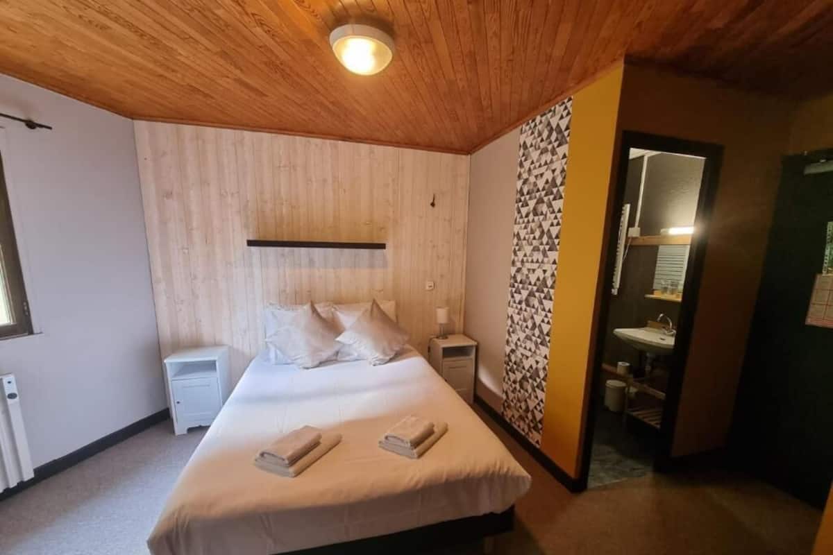 Habitación de hotel con techo de madera, cama con ropa blanca, mesita de noche, lámpara fijada a la pared y baño visible a través de una puerta abierta.