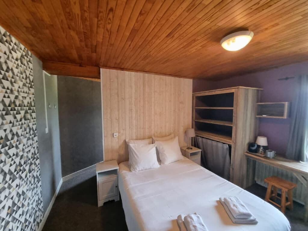 Una habitación de hotel con techo de madera, una cama con ropa de cama blanca, una mesita de noche, un escritorio y una ventana con cortinas.