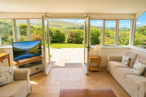 A living room with a TV, a sofa, a wooden side table, and a door leading to a garden. - Claybottom Farm, Caldbeck - in a rural setting with fell walks from the door (Hesket Newmarket)