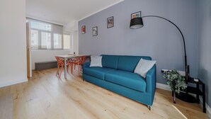 Ein modernes Wohnzimmer mit einem türkisfarbenen Sofa, einem Esstisch mit Stühlen, einer Stehlampe und gerahmten Bildern an der Wand.