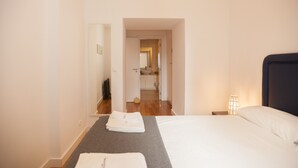Habitación de hotel con una cama, una mesita de noche, una lámpara, una toalla y vistas a un baño y una cocina a través de una puerta abierta.