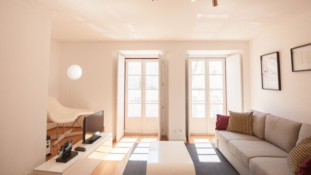 Una sala de estar moderna con sofá, mesa de centro, televisor y una silla. Dos puertas dan acceso a un balcón.