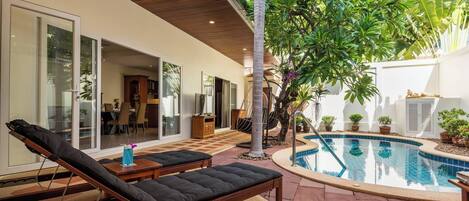 A modern outdoor pool area with lounge chairs, a dining area, and a large tree.