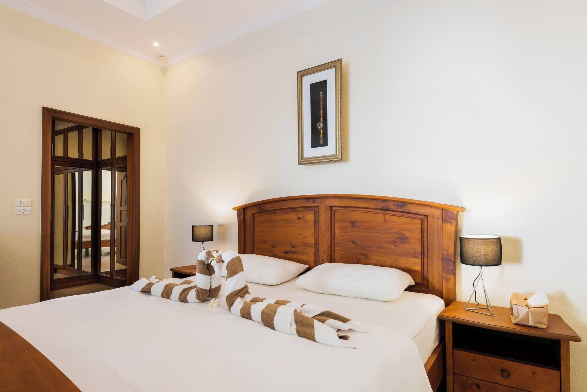 A hotel room with a wooden bed, a nightstand with a lamp, a framed picture on the wall, and a door leading to another room.