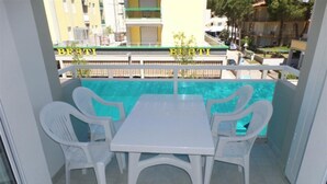 Un balcon avec une table et des chaises blanches, une piscine en arrière-plan et un bâtiment portant l’inscription « BERTI ».