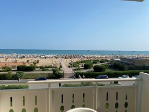 Blick auf einen Strand mit Sonnenschirmen und Menschen, eine Straße mit Autos sowie ein Gebäude mit einem Balkon.