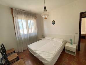 Ein Schlafzimmer mit einem Bett, einem Nachttisch, einem Sessel, einem Fenster mit Vorhängen und einer Wandleuchte.