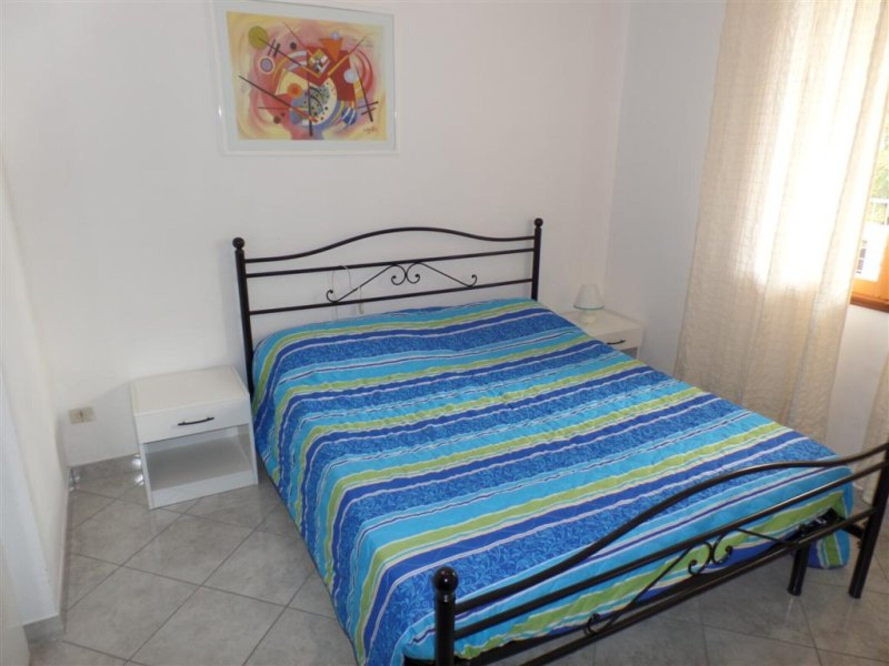 A bedroom with a bed, a bedside table, a nightstand, a drawer, and a painting on the wall.