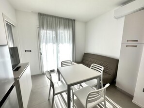 Quarto pequeno e moderno com cozinha compacta, mesa de jantar com cadeiras, sofá e uma janela grande com cortinas.