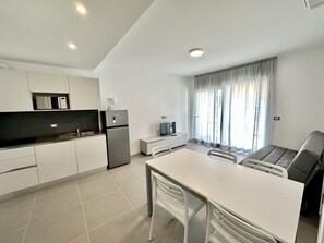 Eine moderne Küche mit weißen Schränken, einem Kühlschrank, einer Mikrowelle und einer Kaffeemaschine. Ein Esstisch mit Stühlen, ein Fernseher und ein Fenster mit Vorhängen sind ebenfalls vorhanden.