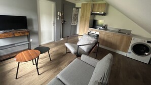 Ein kompakter Wohnraum mit Sofa, Fernseher, kleinem Tisch, Stuhl, Waschmaschine und Küchenzeile.