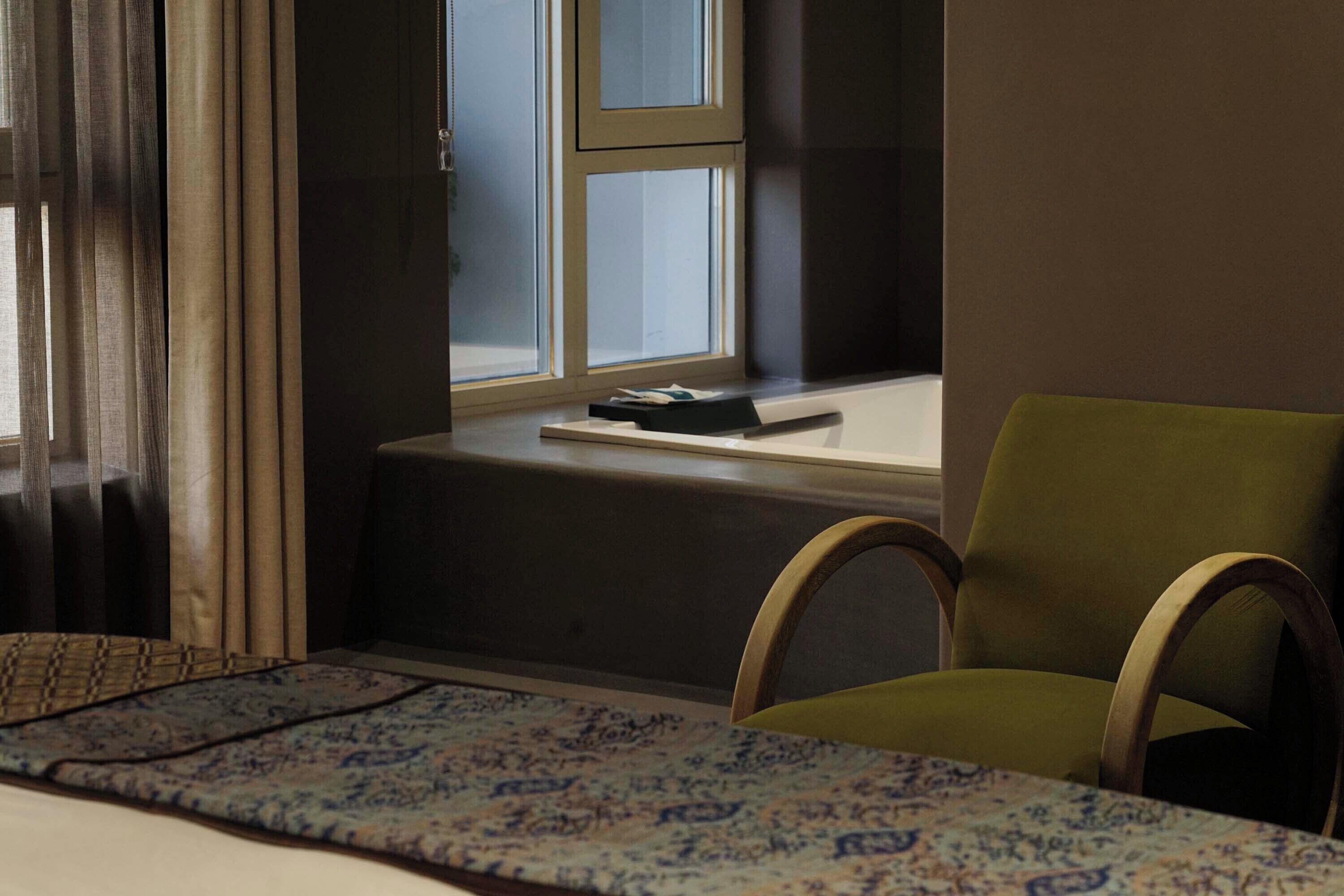 A room with a bed, a window with curtains, a chair, and a small table with a book.