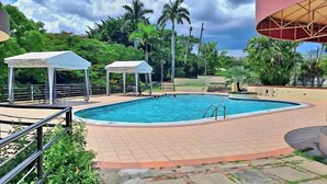 Una piscina con borde de baldosas, rodeada de vegetación y estructuras cubiertas.