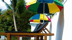 Una silla de lounge de madera con una sombrilla colorida, palmeras y un edificio blanco al fondo.