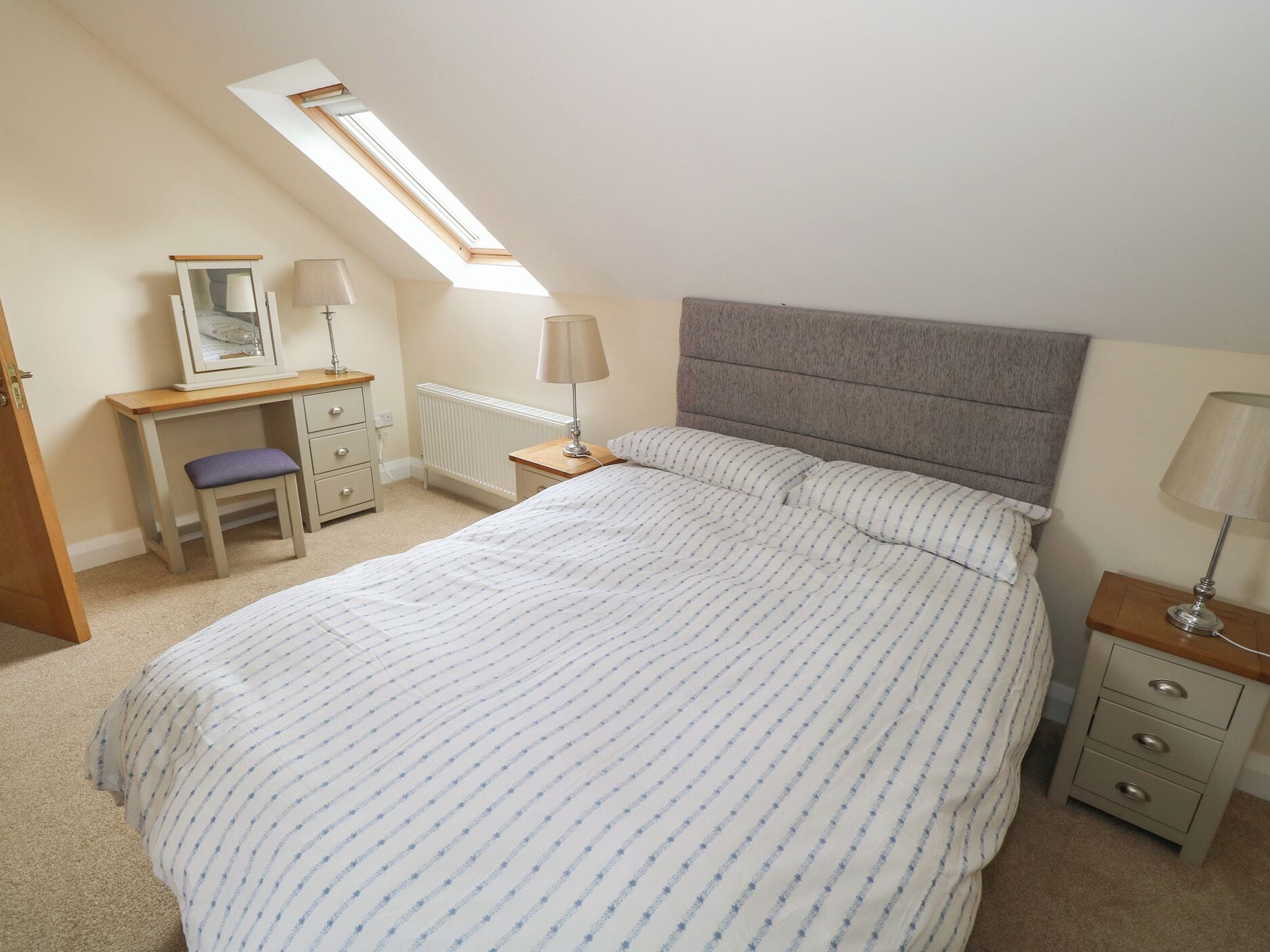 A bedroom with a bed, a desk, a chair, a nightstand, a lamp, a radiator, and a skylight.