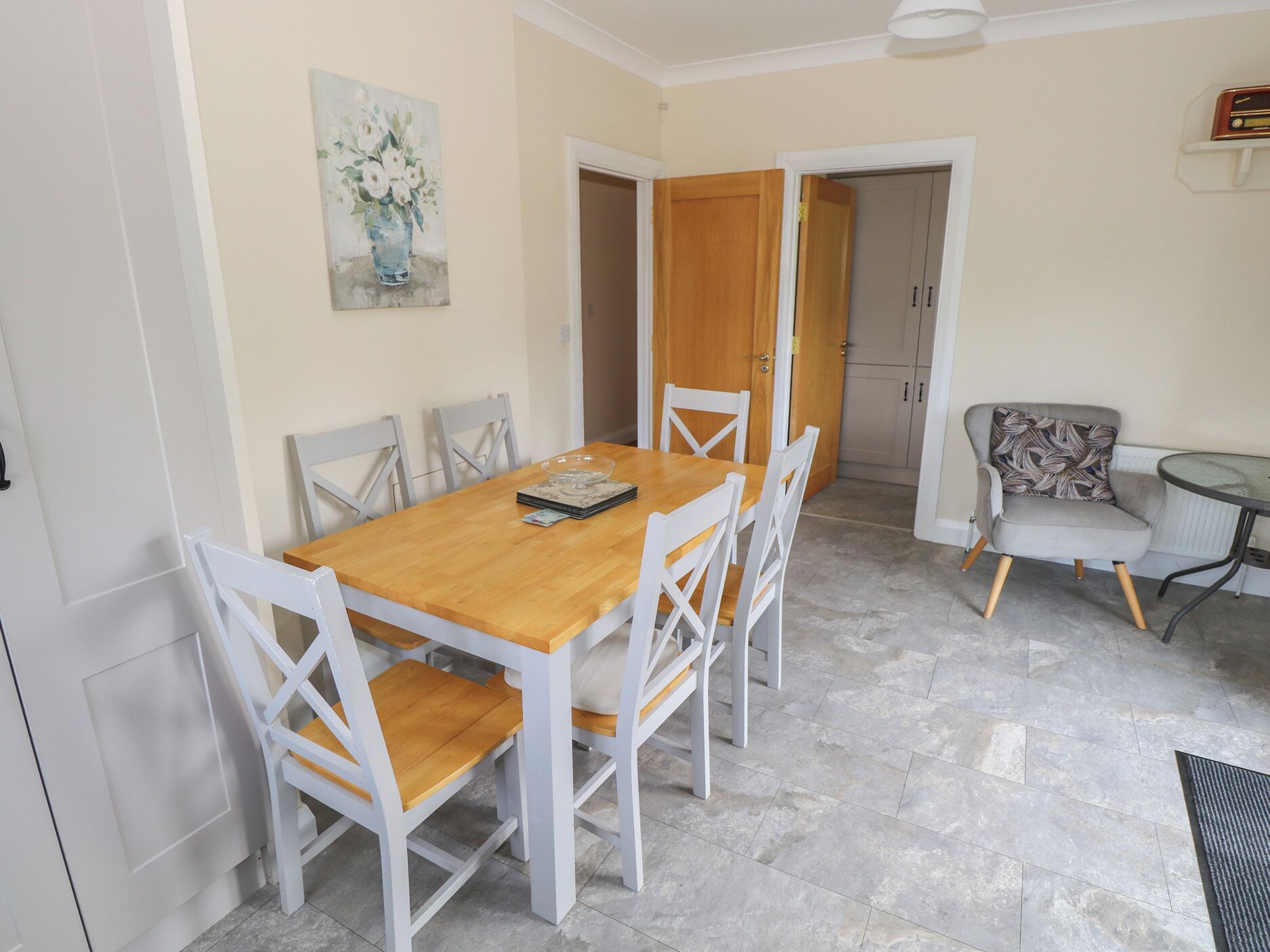 A dining area with a wooden table and white chairs, a small round table with a chair, and a door leading to another room.