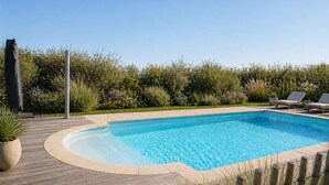 Una piscina rettangolare con un bordo in legno, due lettini da relax e vario verde sullo sfondo.