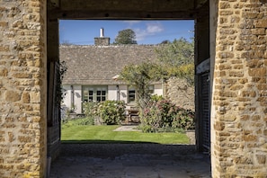 Un arco de piedra que conduce a un edificio con techo de tejas, una chimenea y un jardín con flores y un banco.