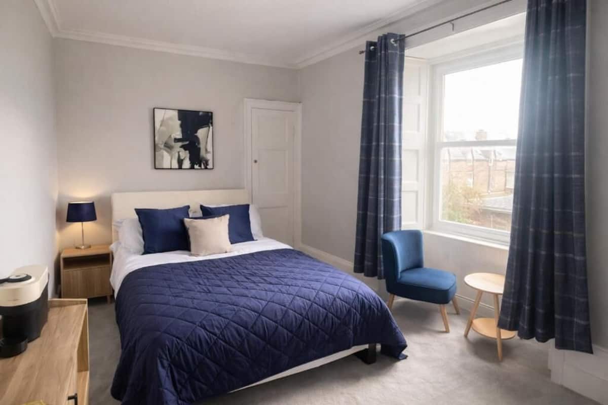 A bedroom with a bed, a chair, a small table, a lamp, and a window with curtains.