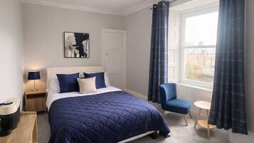 A bedroom with a bed, a chair, a small table, a nightstand, a lamp, a painting, and a window with curtains.