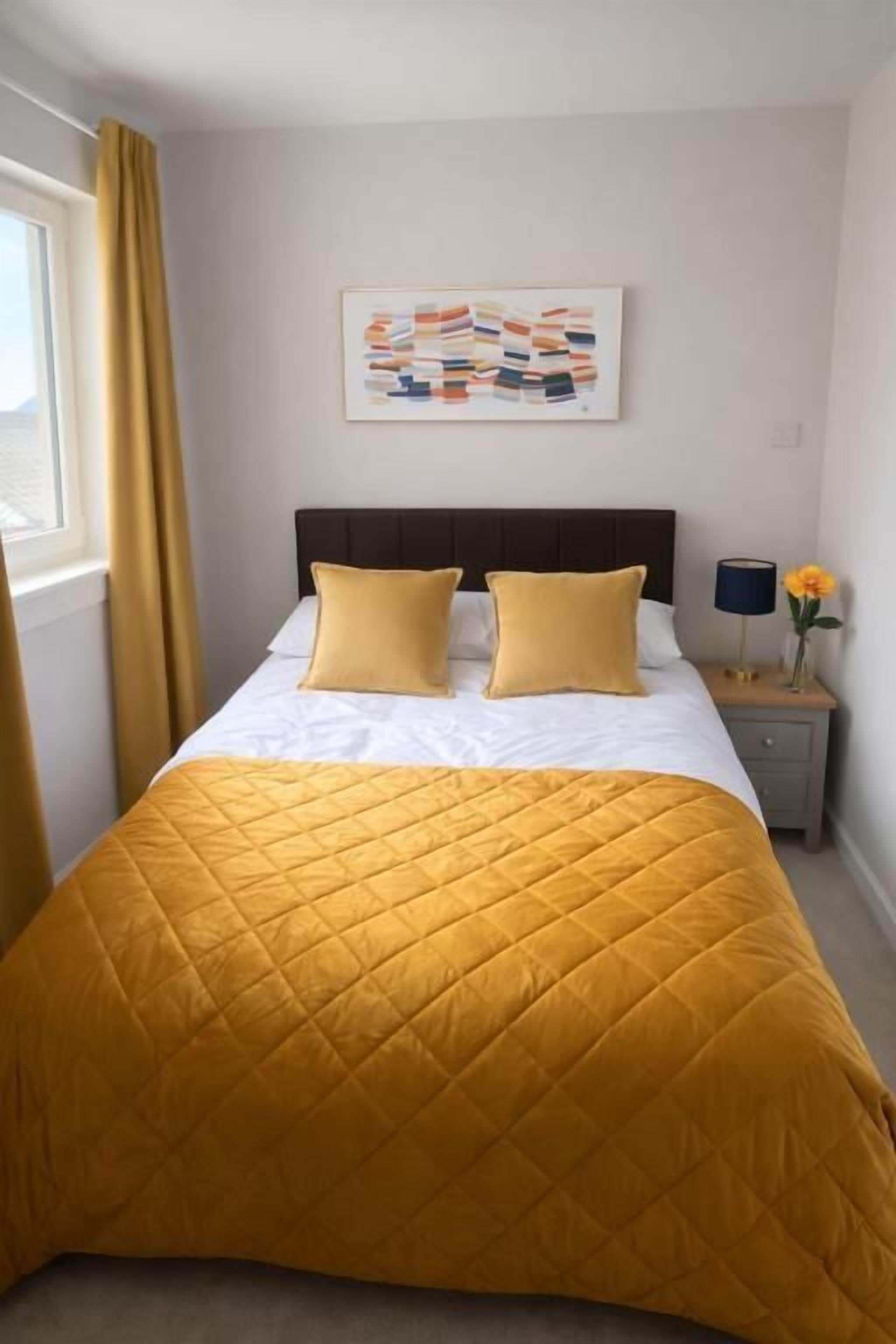 A bed with a quilted yellow bedspread, a white headboard, and two pillows. There is a framed artwork on the wall above the bed and a bedside table with a lamp and a vase with flowers.
