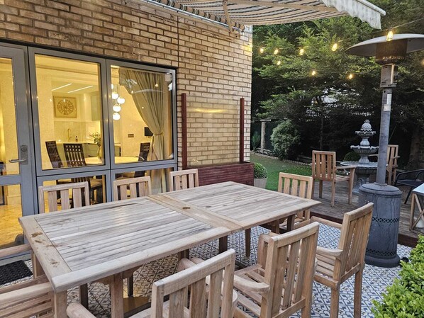 Eine Terrasse mit Holztischen und -stühlen, eine Ziegelwand und ein Garten mit einem Brunnen.