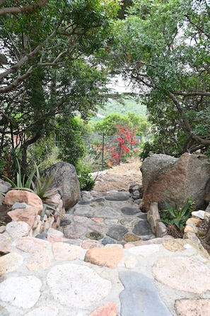 A stone pathway leading through a garden with large rocks, various plants, and a view of a hillside. - Two doubles with beautiful sea views in Costa Rei (Costa Rei)