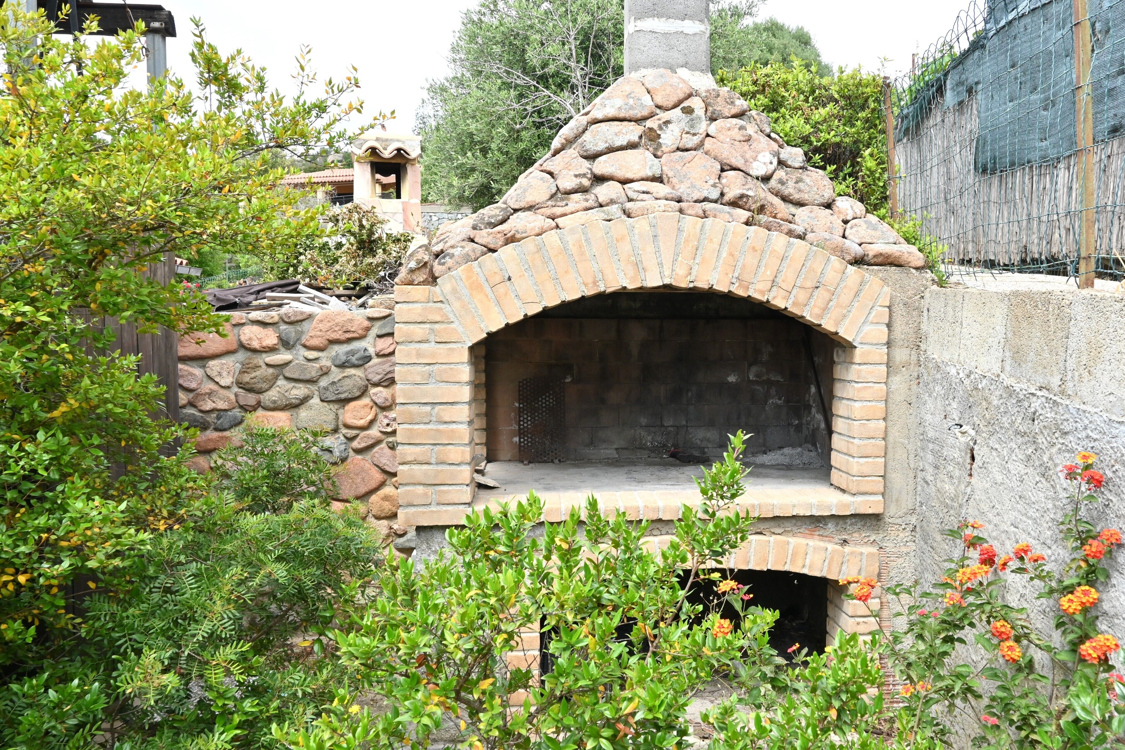 Un horno de piedra al aire libre con chimenea de ladrillo, rodeado de vegetación y un muro de piedra.