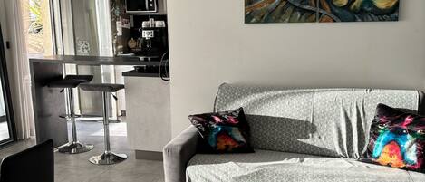Una sala de estar moderna con un sofá gris, almohadones coloridos, un cuadro en la pared y una cocina al fondo.