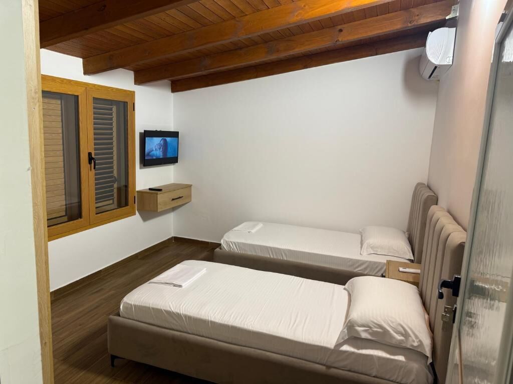 A room with two beds, a TV, a wooden ceiling, and a window with shutters.