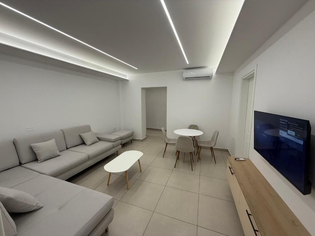 A modern living room with a sofa, a small table, a dining area, and a TV mounted on the wall.
