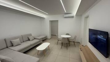 A modern living room with a sofa, a small table, a dining area, and a TV mounted on the wall.