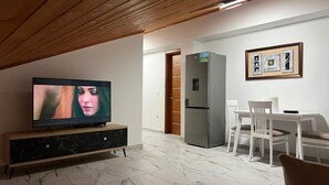 Una sala de estar con televisor, un comedor con mesa y sillas, refrigerador y techo de madera.