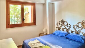 A bedroom with a bed, a window with a view of greenery, a bedside table with a lamp, and a decorative metal headboard. - Villino Marzamemi (Marzamemi)