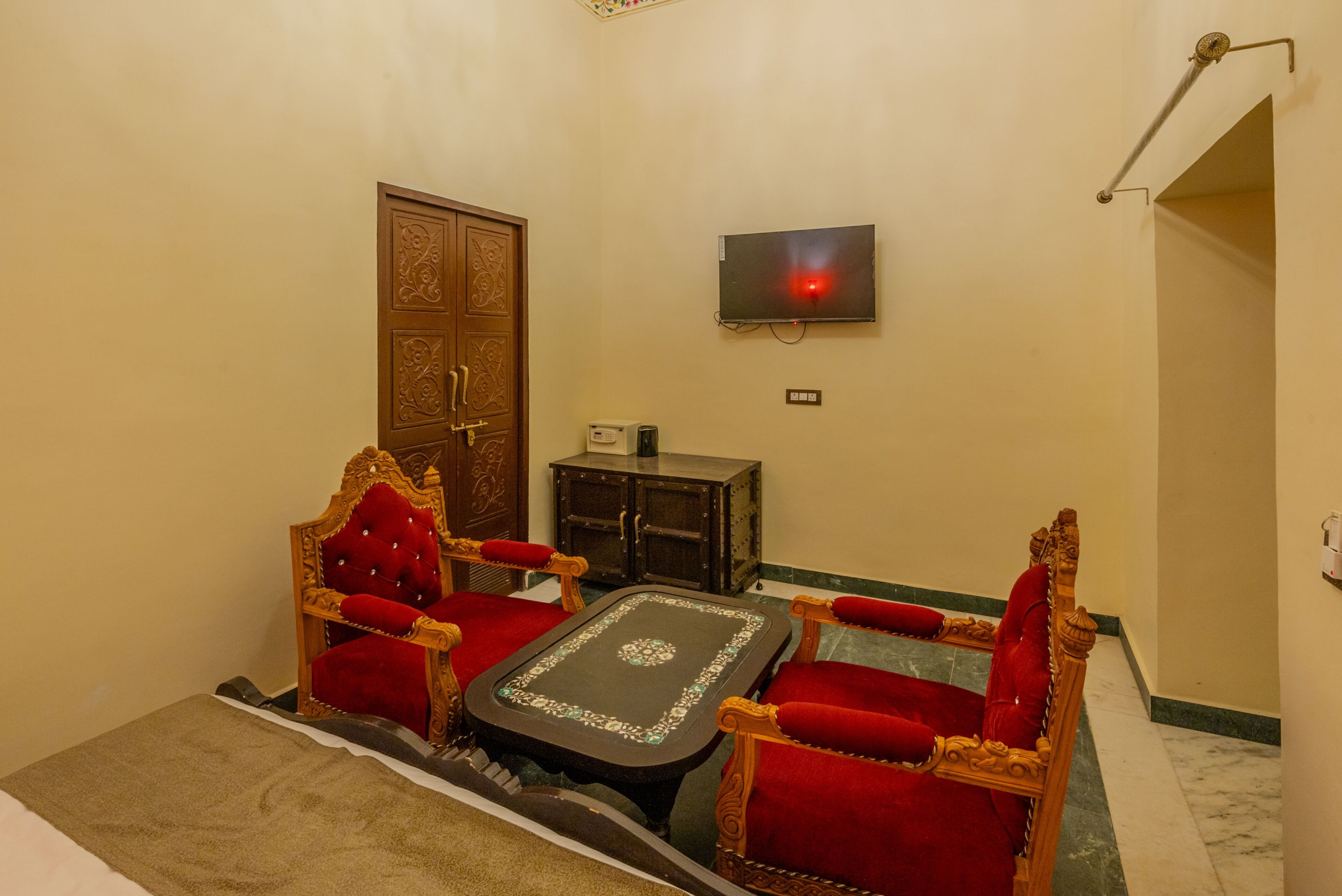 A room with a wooden door, a television mounted on the wall, a small cabinet, and two red upholstered chairs with a matching table.