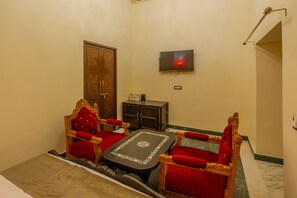 A room with a wooden door, a television mounted on the wall, a small cabinet, and two red upholstered chairs with a matching table. - Mahal Khas Palace (Bharatpur)