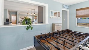 A room with a foosball table, a window with a view, and a kitchen area visible through an open door. - Sea Salt & Sunny Days: Pool, Spa, Putting Green by Anna Maria Life Vacation Rentals (Holmes Beach)