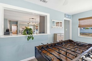 A room with a foosball table, a window with a view, and a kitchen area visible through an open door. - Sea Salt & Sunny Days: Pool, Spa, Putting Green by Anna Maria Life Vacation Rentals (Holmes Beach)