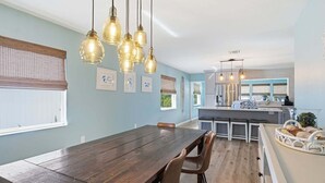 A dining area with a wooden table and chairs, a kitchen with a central island, and pendant lighting. - Sea Salt & Sunny Days: Pool, Spa, Putting Green by Anna Maria Life Vacation Rentals (Holmes Beach)