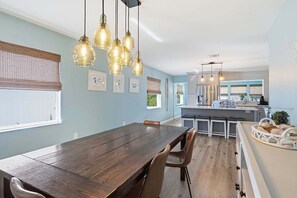 A dining area with a wooden table and chairs, a kitchen with a central island, and pendant lighting. - Sea Salt & Sunny Days: Pool, Spa, Putting Green by Anna Maria Life Vacation Rentals (Holmes Beach)